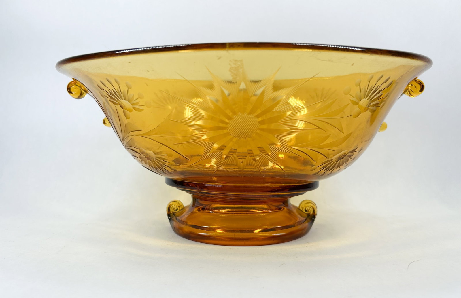 VINTAGE AMBER GLASS FOOTED BOWL: A vintage amber glass footed bowl featuring a molded floral and starburst pattern. The vessel is designed with two applied scroll handles and sits on a pedestal base with three scrolled feet.
