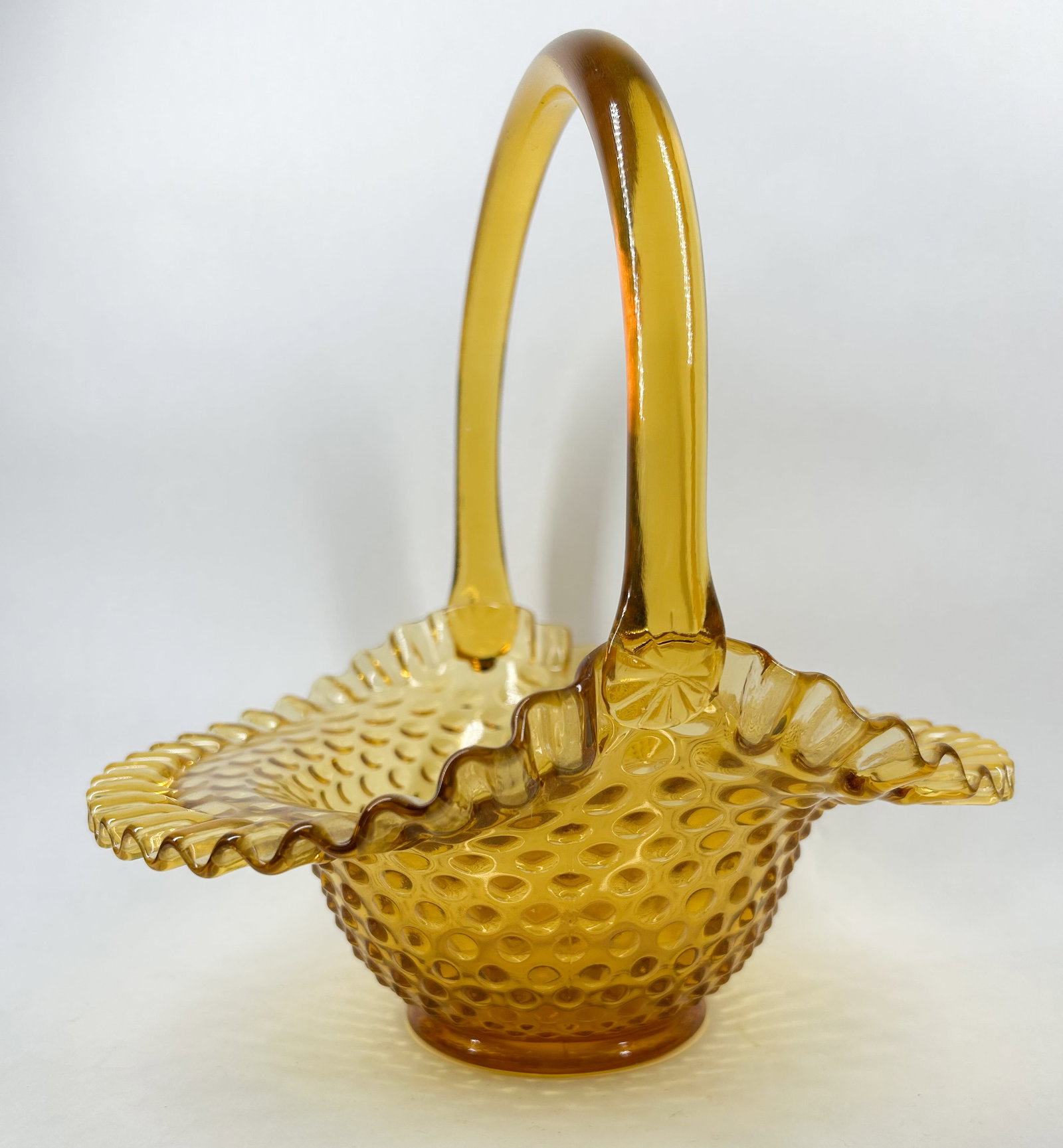 AMBER HOBNAIL GLASS BASKET, 20TH CENTURY: Vintage transparent amber glass basket featuring a molded hobnail pattern and a deeply crimped, ruffled rim. The piece is constructed with a high arched handle applied with molded rosette joins and