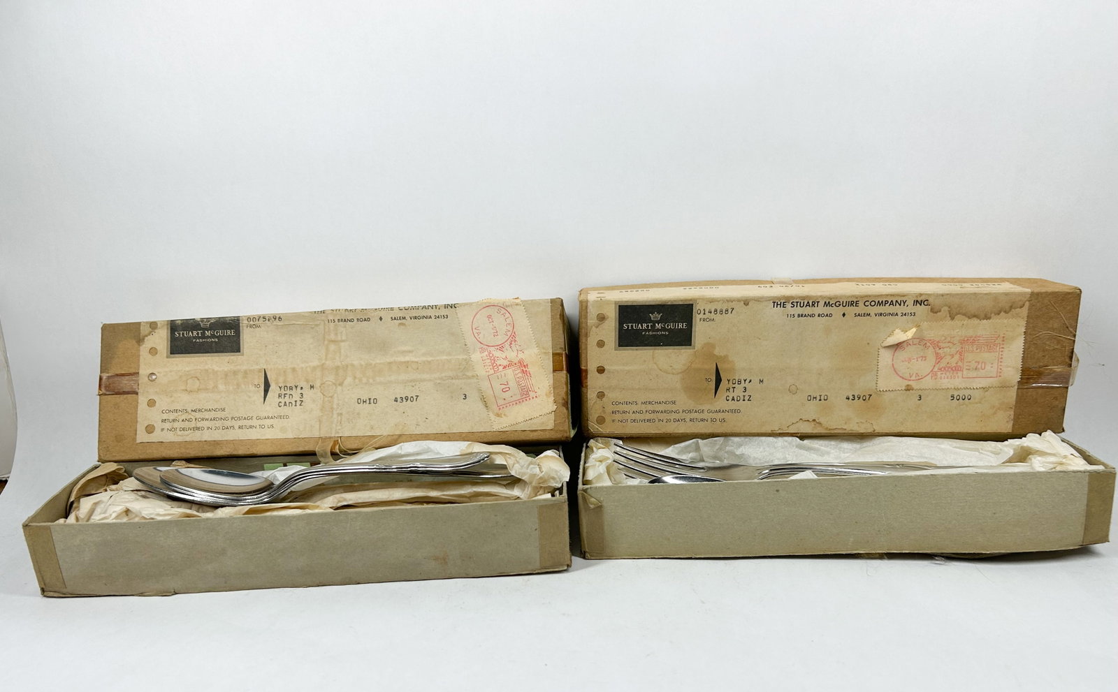 STUART MCGUIRE STAINLESS FLATWARE, 1970S: Two sets of Stuart McGuire stainless steel flatware housed in their original shipping boxes. Each box bears a printed label reading STUART McGUIRE FASHIONS with a crown logo and remains addressed to C