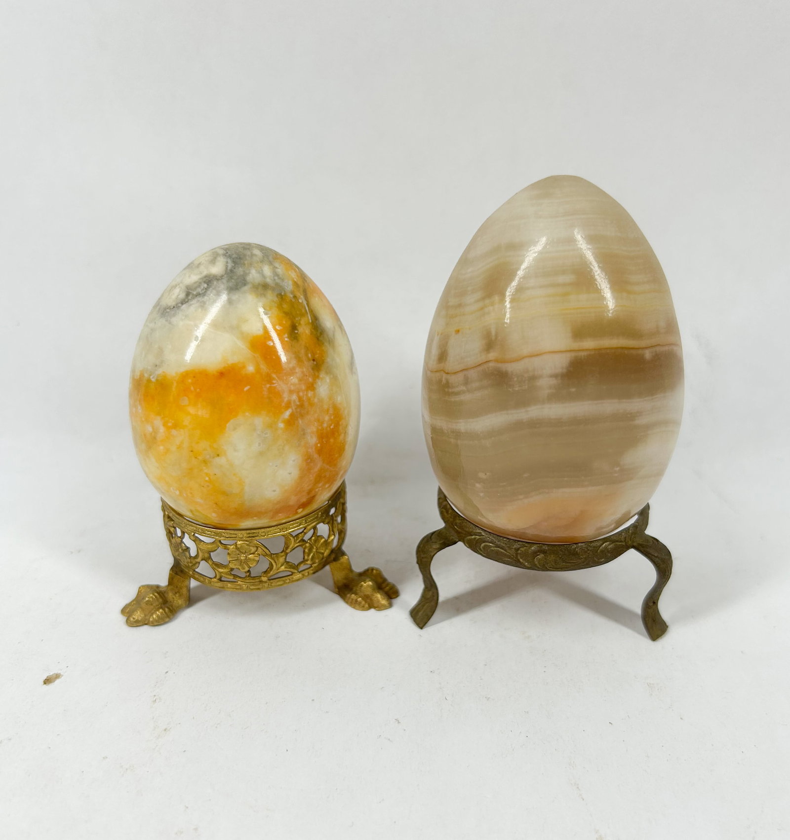 TWO POLISHED ONYX DECORATIVE EGGS, 20TH CENTURY: Two polished stone decorative eggs, one featuring mottled yellow and orange hues and the other displaying banded cream and tan layers. Each specimen rests upon an individual cast brass-toned