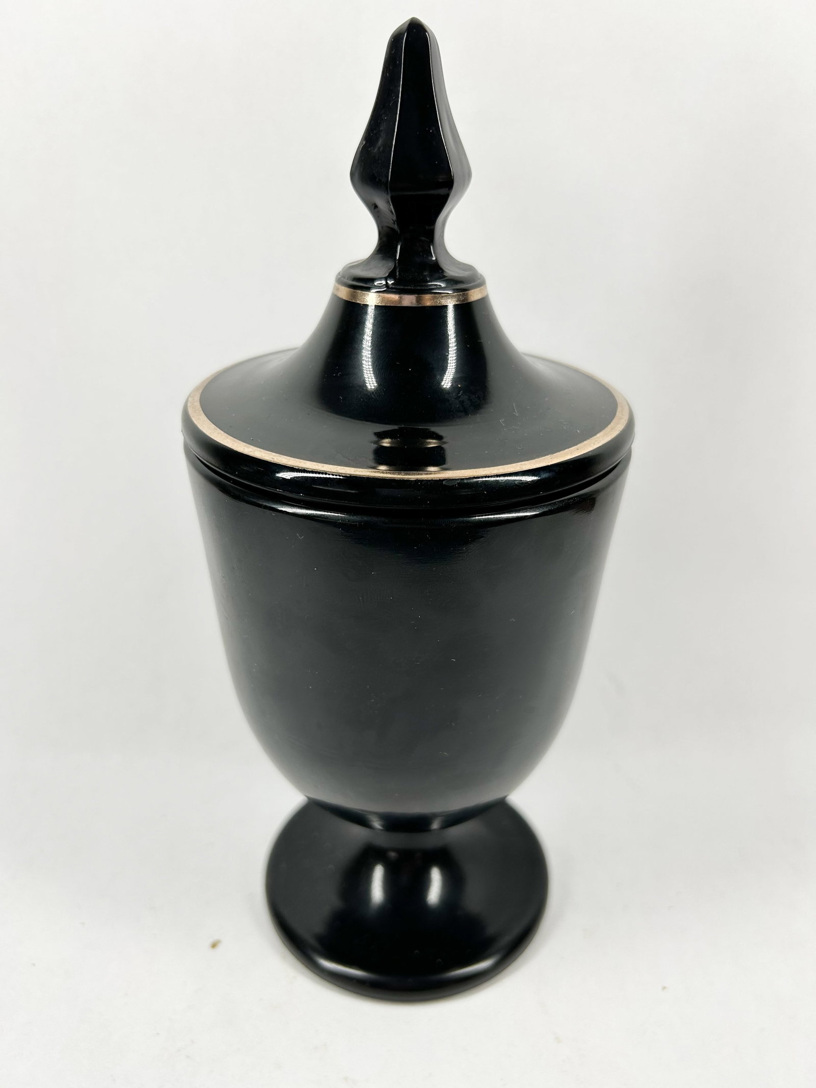 BLACK GLASS LIDDED TROPHY URN, FENTON: Opaque black glass lidded trophy urn featuring silver-toned metallic banding. The vessel is designed with a tapered body on a flared pedestal foot, paired with a matching domed lid and faceted