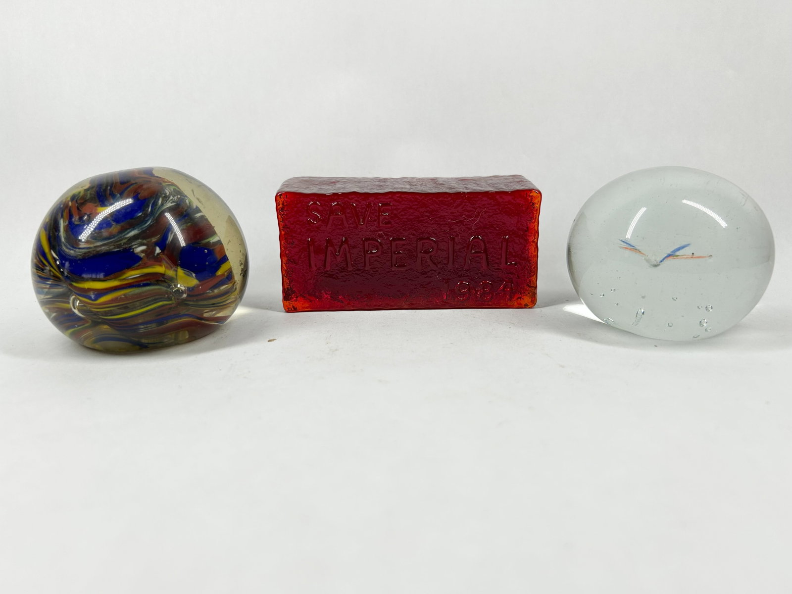 IMPERIAL GLASS BRICK AND PAPERWEIGHTS, 1984: Imperial Glass Corporation ruby red commemorative brick and two art glass paperweights. The brick is embossed with SAVE IMPERIAL 1984, and the paperweights feature an internal multi-color swirl and a