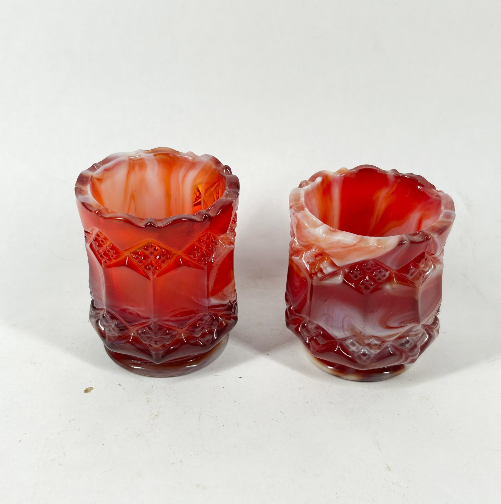 IMPERIAL RED SLAG GLASS TOOTHPICK HOLDERS: A pair of pressed slag glass toothpick holders featuring a marbled red and white colorway. The vessels are decorated with a central band of embossed diamond motifs above a textured base.