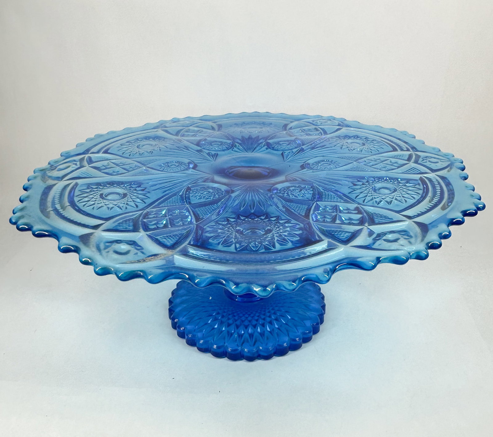 Cobalt Blue Pinwheel and Star Pattern Glass Cake Stand: A pressed cobalt blue glass pedestal cake stand featuring the Pinwheel and Star pattern. The flat, circular plate is decorated with a radial design composed of six large pinwheels interspersed with