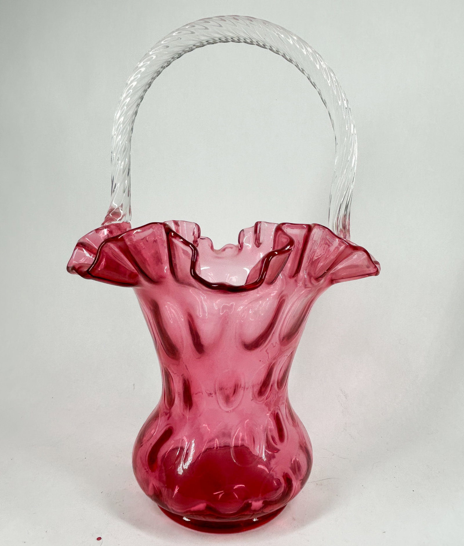 FENTON CRANBERRY THUMBPRINT GLASS BASKET, 1995: A cranberry glass basket featuring a thumbprint pattern, a ruffled rim, and a clear twisted glass handle. The base is marked with a frosted oval stamp reading FENTON and 90th ANNIVERSARY, identifying