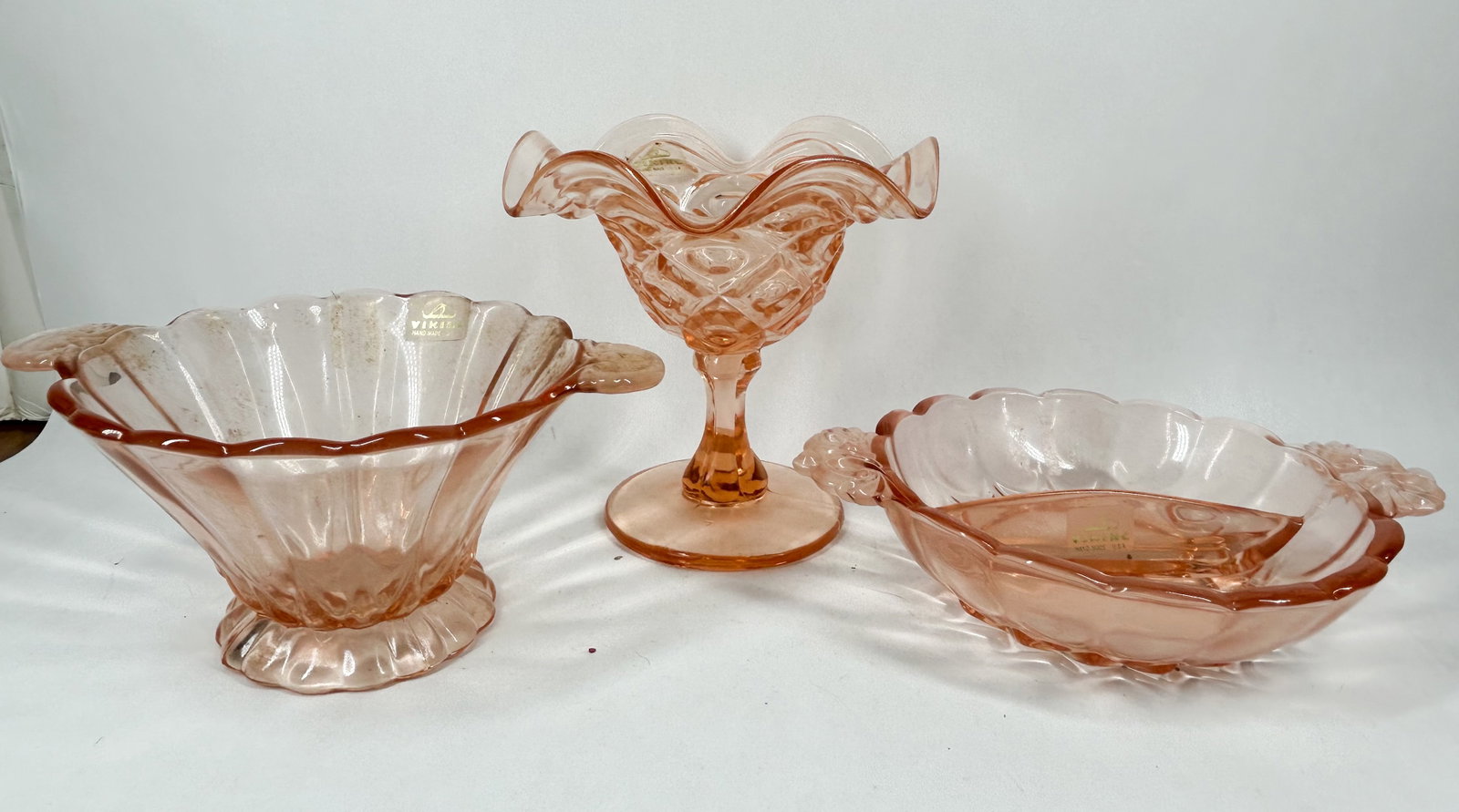 VIKING PINK GLASS TABLEWARE TRIO, C.1970: Viking Glass translucent pink serving set comprising a handled bowl, ruffled compote, and divided dish. Each item features a rectangular gold adhesive label printed with a sailboat logo and text