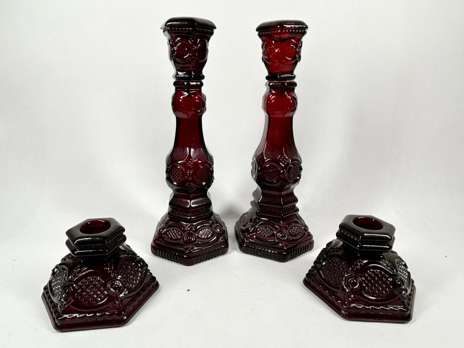 AVON CAPE COD RUBY GLASS, 1970S: A set of four ruby red pressed glass items from the 1876 Cape Cod Collection produced by Avon Products, Inc. One tall piece retains its original circular paper label for Bird of Paradise Cologne, and