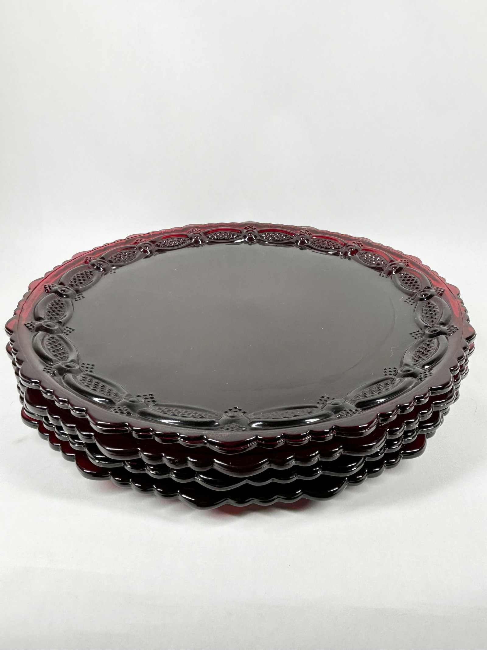 AVON CAPE COD RUBY RED GLASS PLATES: A set of pressed glass plates in the 1876 Cape Cod pattern by Avon. Each plate features a decorative rim with an interlocking oval motif and a sunburst pattern at the center. The name AVON is