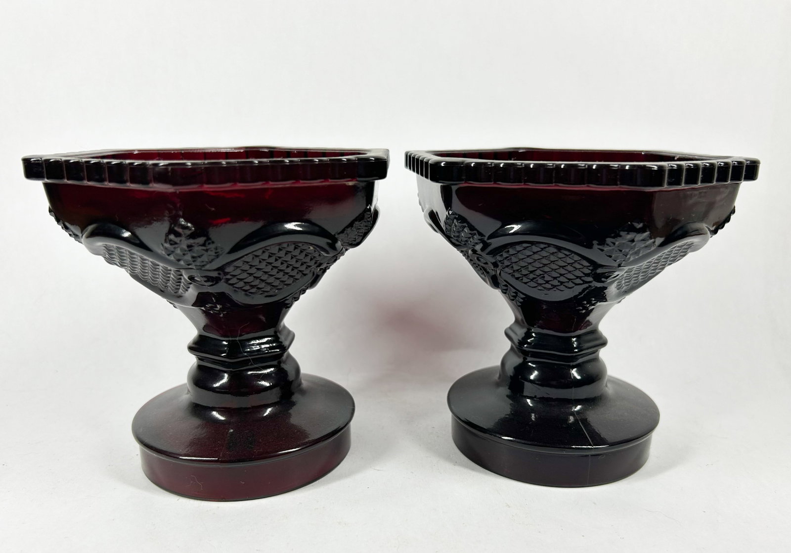 PAIR OF RUBY RED GLASS COMPOTES, MID-CENTURY: Pair of mid-20th-century footed compotes in deep ruby red pressed glass. The hexagonal basins feature scalloped rims and embossed decorative medallions above tiered pedestal bases with visible mold