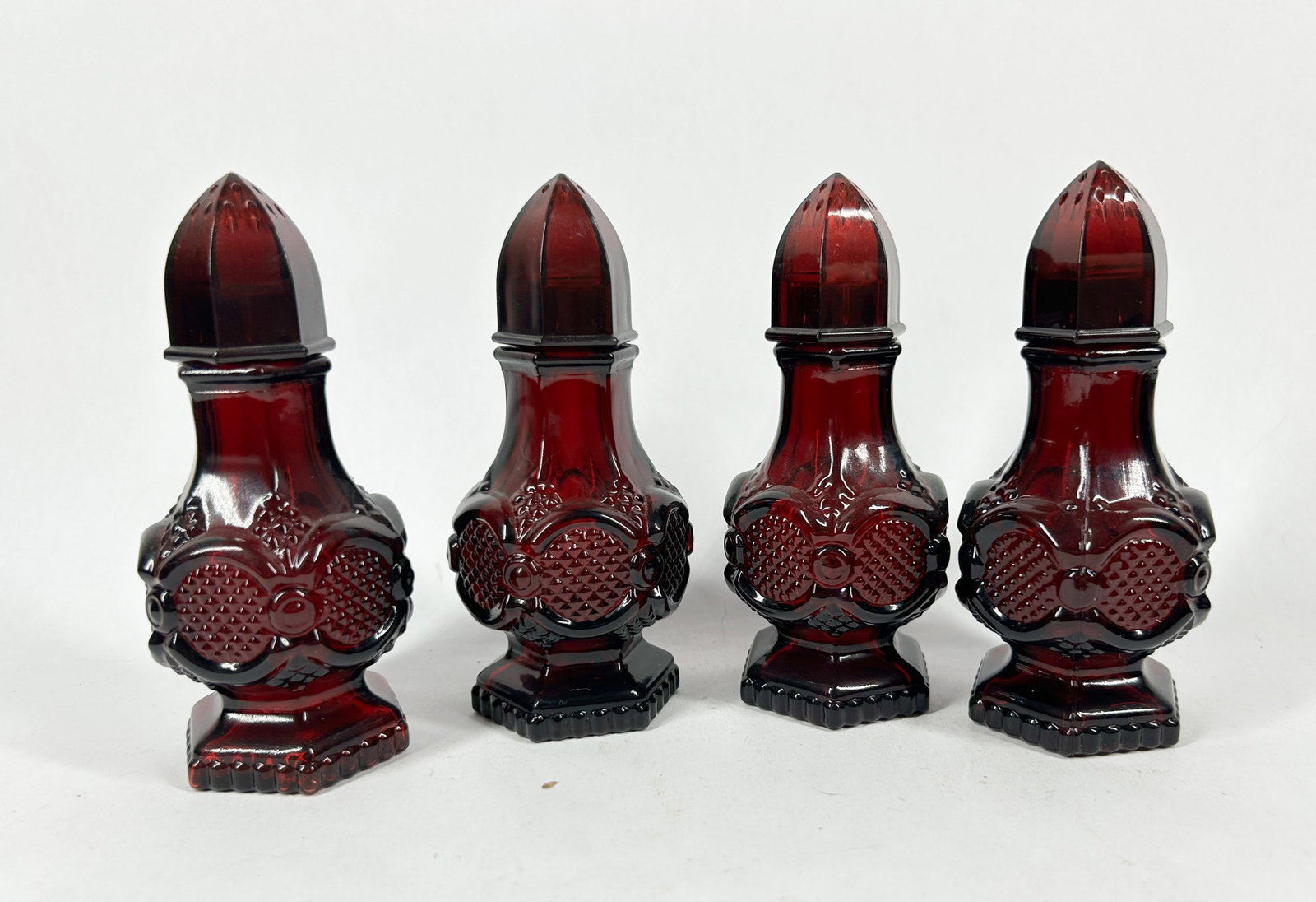AVON RUBY GLASS COLOGNE SET 1970S: A set of four ruby red pressed glass cologne bottles produced by Avon Products, Inc. in the form of salt shakers with faceted plastic caps. Each base retains a white adhesive label reading SALT