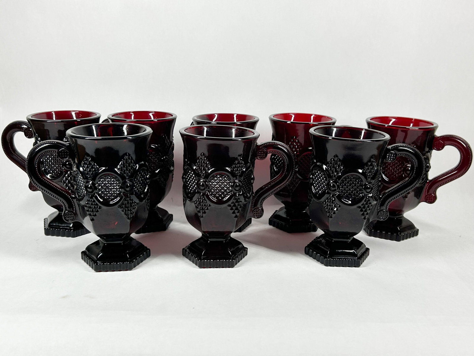 AVON RUBY GLASS MUGS, CIRCA 1975: A set of Avon ruby red pressed glass pedestal mugs in the 1876 Cape Cod pattern. The underside of the base features an embossed mark reading AVON followed by a copyright symbol and the number 64.