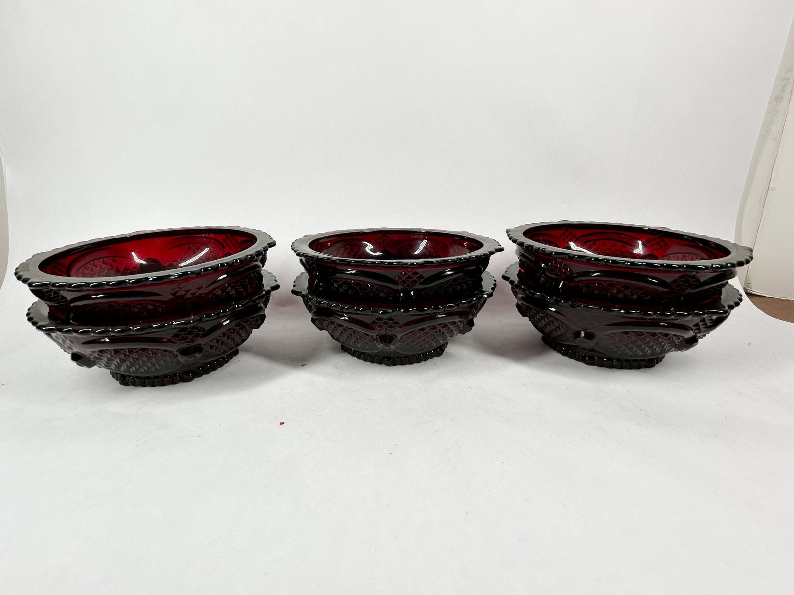 AVON RUBY RED GLASS BOWLS: A collection of Avon 1876 Cape Cod Collection dessert bowls in ruby red pressed glass. Each bowl features a textured diamond-lattice exterior with a scalloped, beaded rim and bears an embossed mark