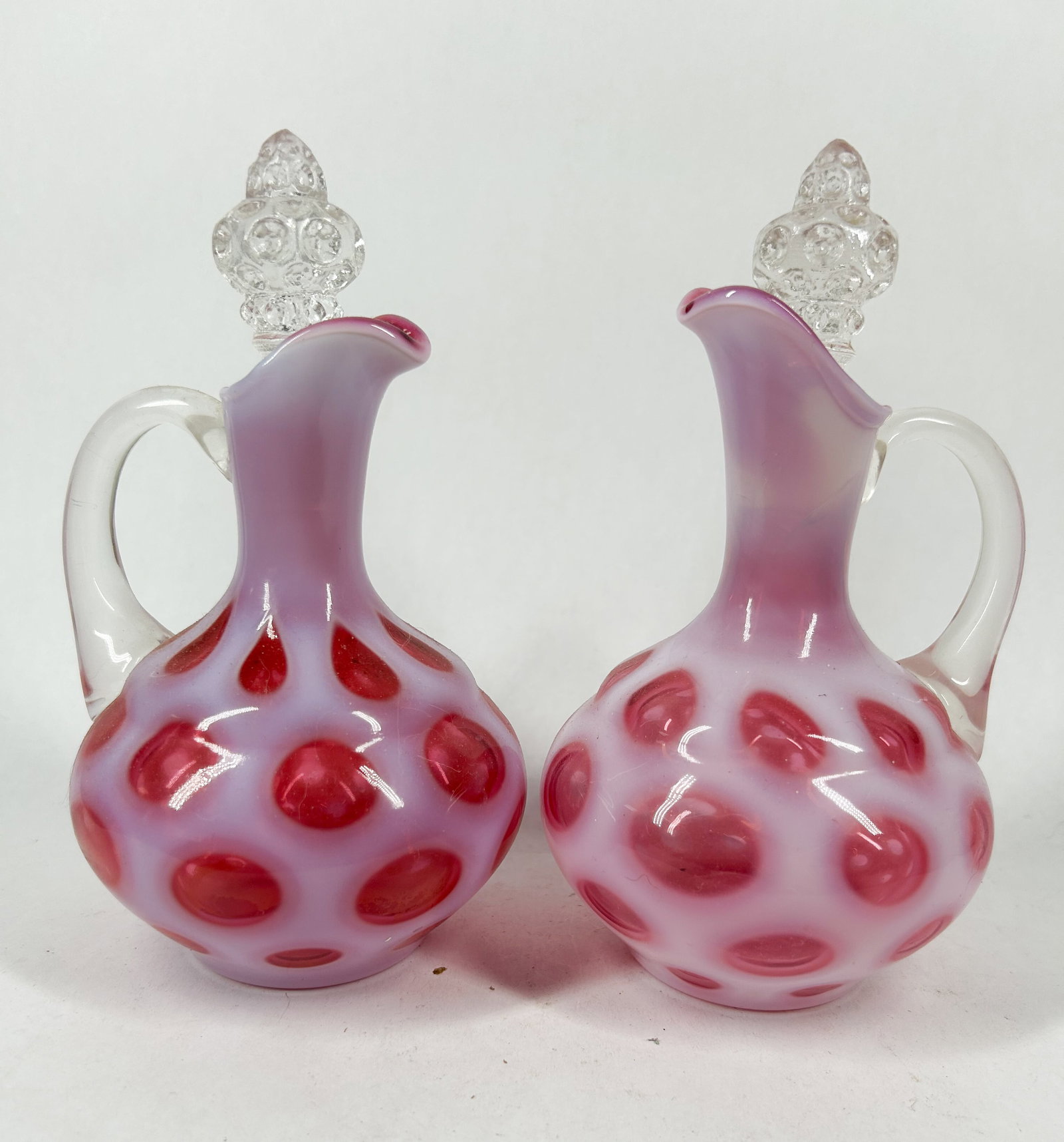 FENTON CRANBERRY OPALESCENT COIN DOT GLASS COLLECTION: An assortment of cranberry opalescent glass vessels featuring a coin dot pattern, including cruets with clear stoppers, a pedestal vase, and rose bowls. A rectangular white adhesive label on the base