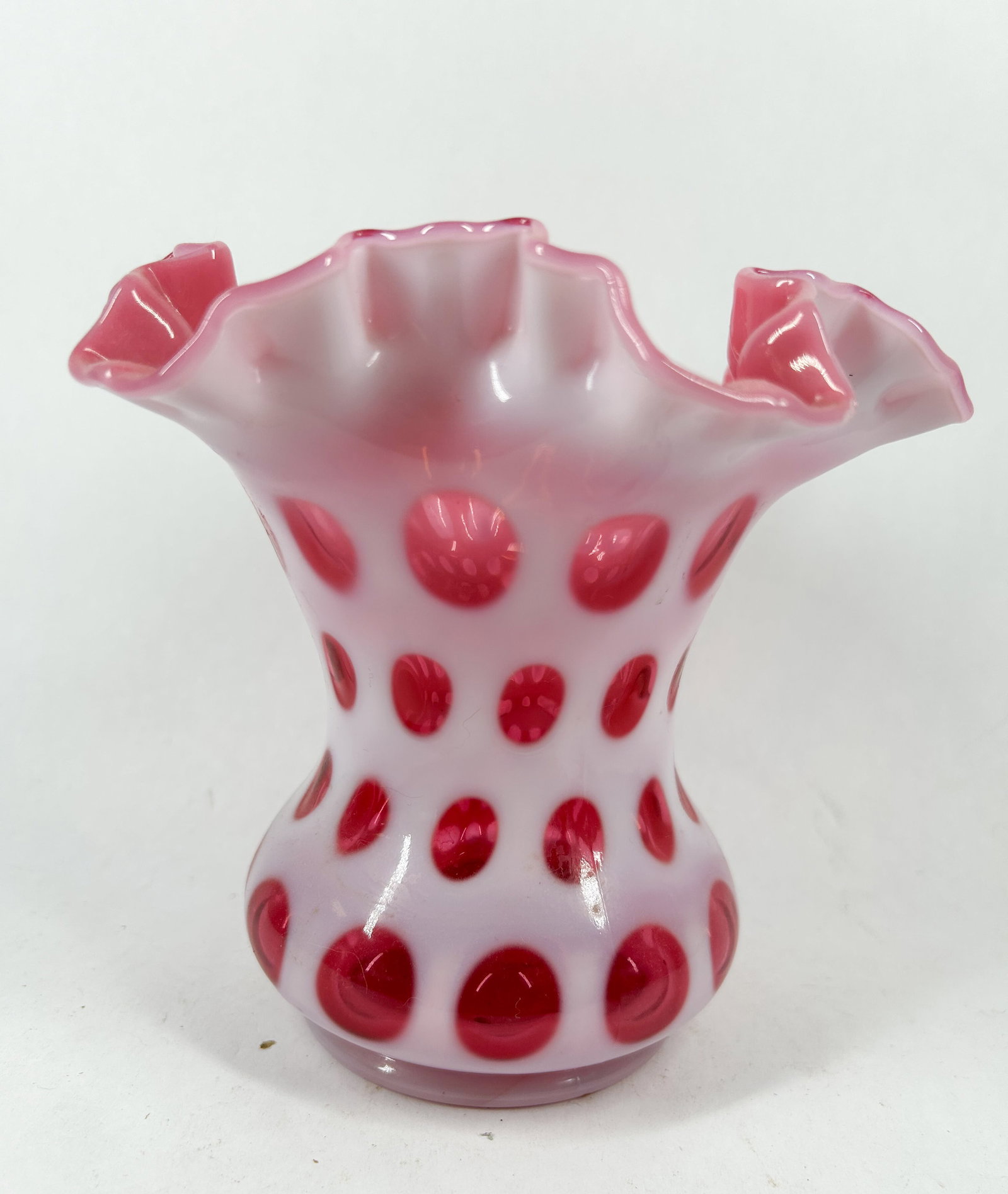 FENTON CRANBERRY OPALESCENT COIN DOT VASE: A mid-20th century cased glass vase featuring a cranberry interior and a white opalescent coin spot pattern. The vessel is designed with a wide ruffled rim and a bulbous body.