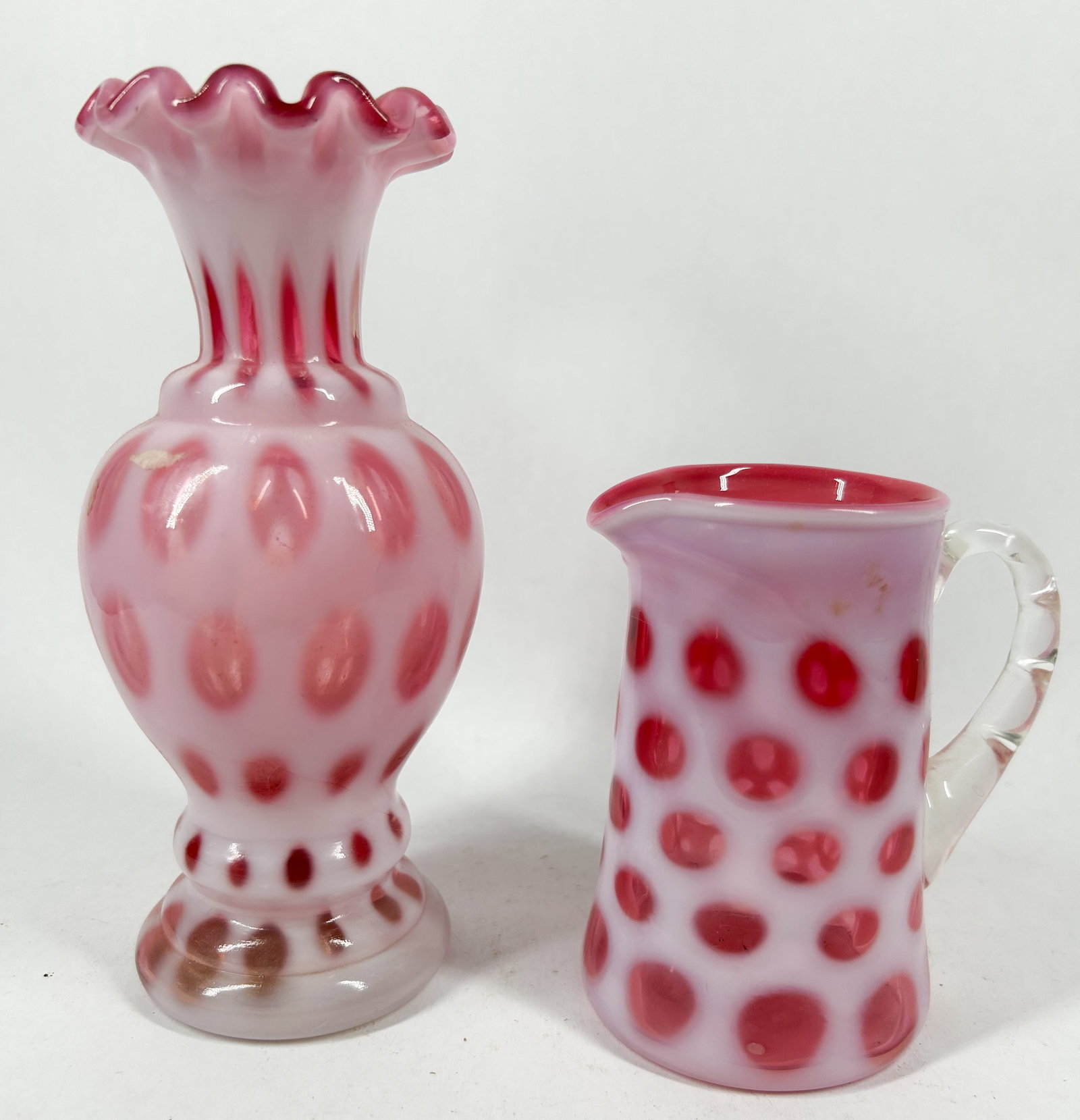 FENTON CRANBERRY OPALESCENT COIN DOT GLASSWARE: Two cranberry opalescent glass vessels comprising a footed ruffled vase and a pitcher with an applied clear reeded handle. The translucent pink bodies are decorated with white opalescent spots, and