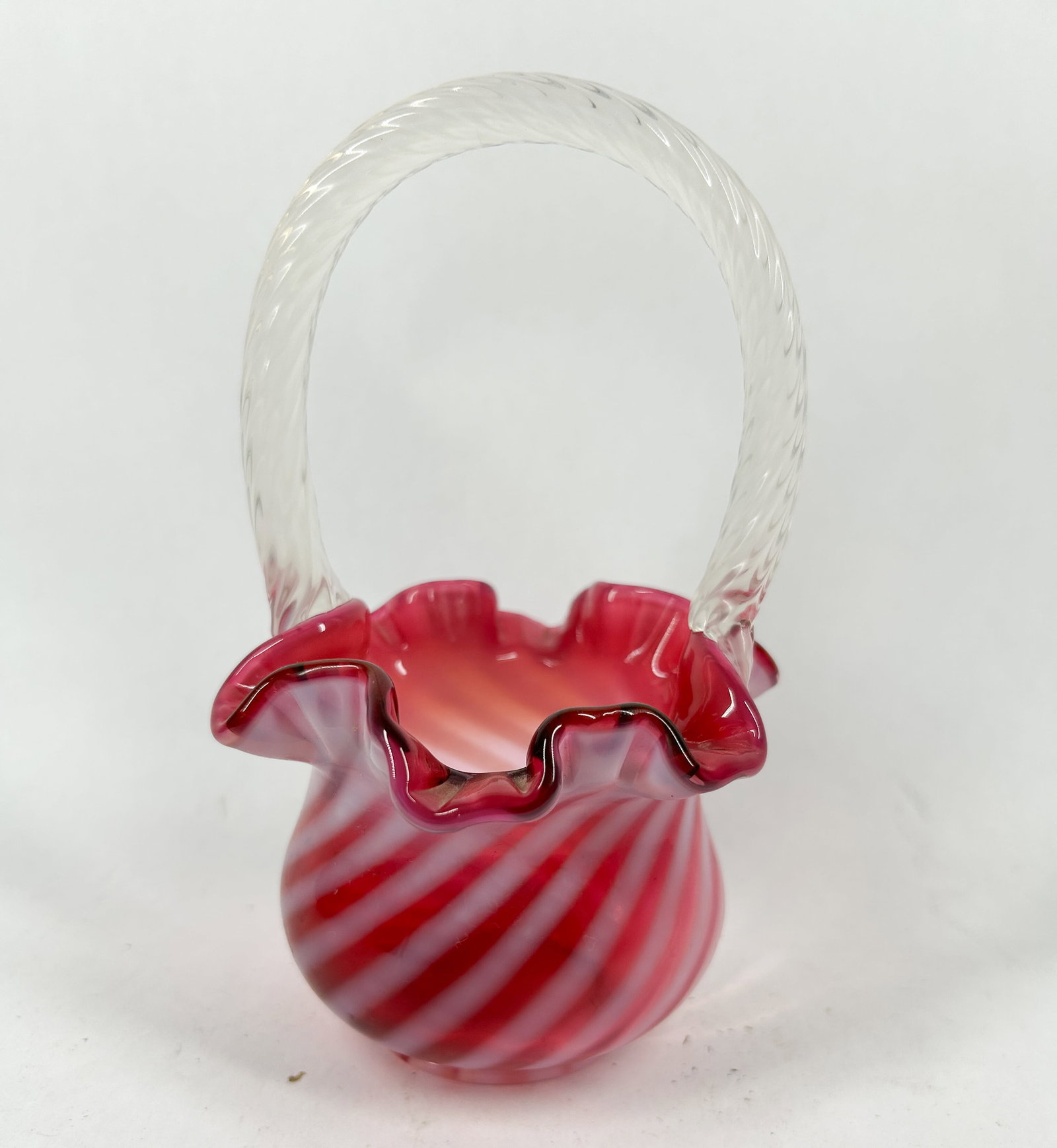 FENTON CRANBERRY OPALESCENT GLASS BASKET, 1970S: Cranberry opalescent glass basket featuring a globular body with diagonal white swirls and a crimped rim, topped with an applied clear twisted glass handle. An impressed oval mark reading Fenton in