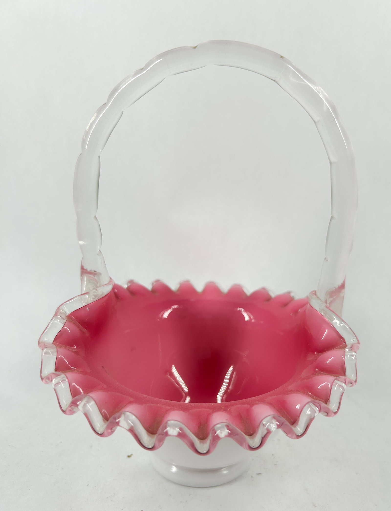 FENTON CASED PINK AND WHITE GLASS BASKET: A rose-pink and white cased glass basket. The vessel features a flared ruffled rim with a clear glass crest and an applied clear glass handle with a molded segmented pattern.