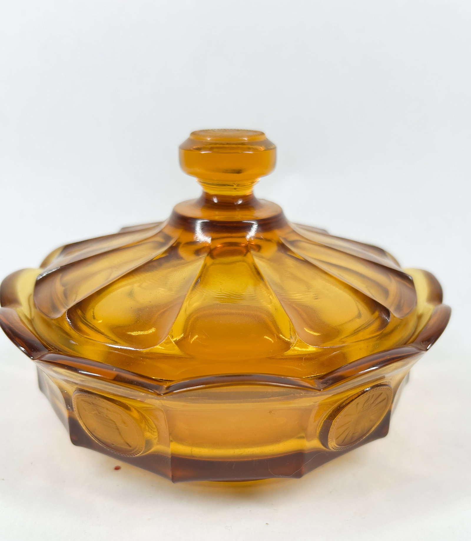 FOSTORIA AMBER GLASS CANDY DISH, C.1960: A lidded honey-amber glass candy dish in the 'Coin' pattern. The circular bowl features a scalloped rim and a series of frosted medallions depicting various motifs including the Liberty Bell and a