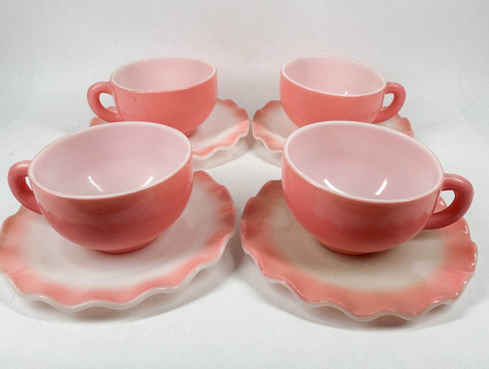 HAZEL ATLAS RUFFLED TABLEWARE: A set of four cups and four saucers in the Ripple pattern featuring a fired-on pink glaze and opaque white glass interiors. The underside of each cup is embossed with a monogram of the letter H over