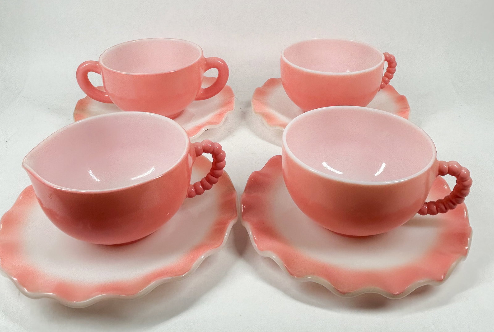HAZEL-ATLAS PINK MILK GLASS SET, C. 1950S: Hazel-Atlas Glass Company tableware set in pink milk glass featuring the Crinoline pattern with ruffled edges and beaded handles. The underside of the bases bears an impressed monogram consisting of