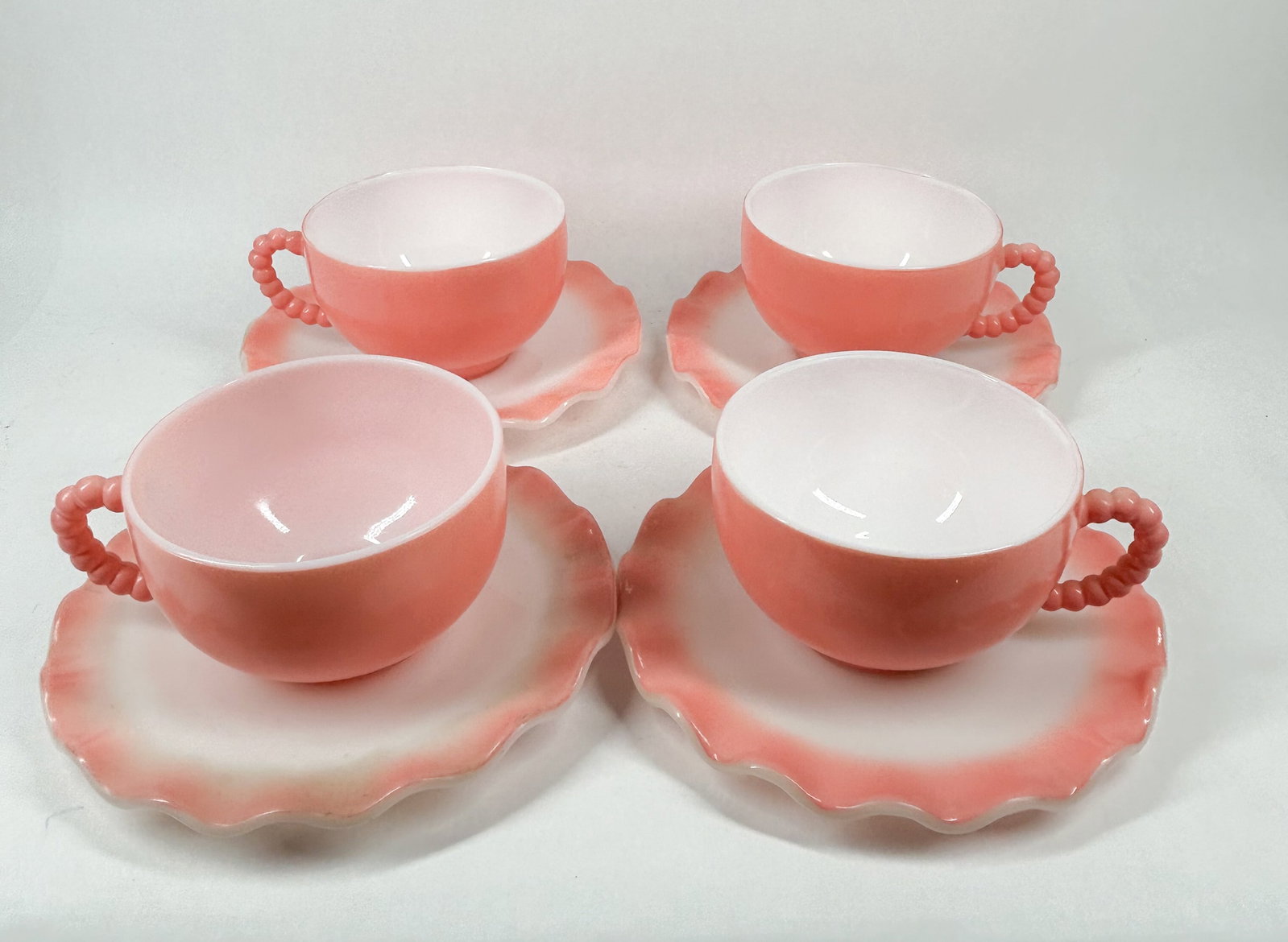 HAZEL ATLAS MILK GLASS TEA SERVICE: A set of pink-shaded milk glass teacups and saucers featuring ruffled edges and beaded handles. An impressed monogram is visible to the center of a saucer underside (photo 8).