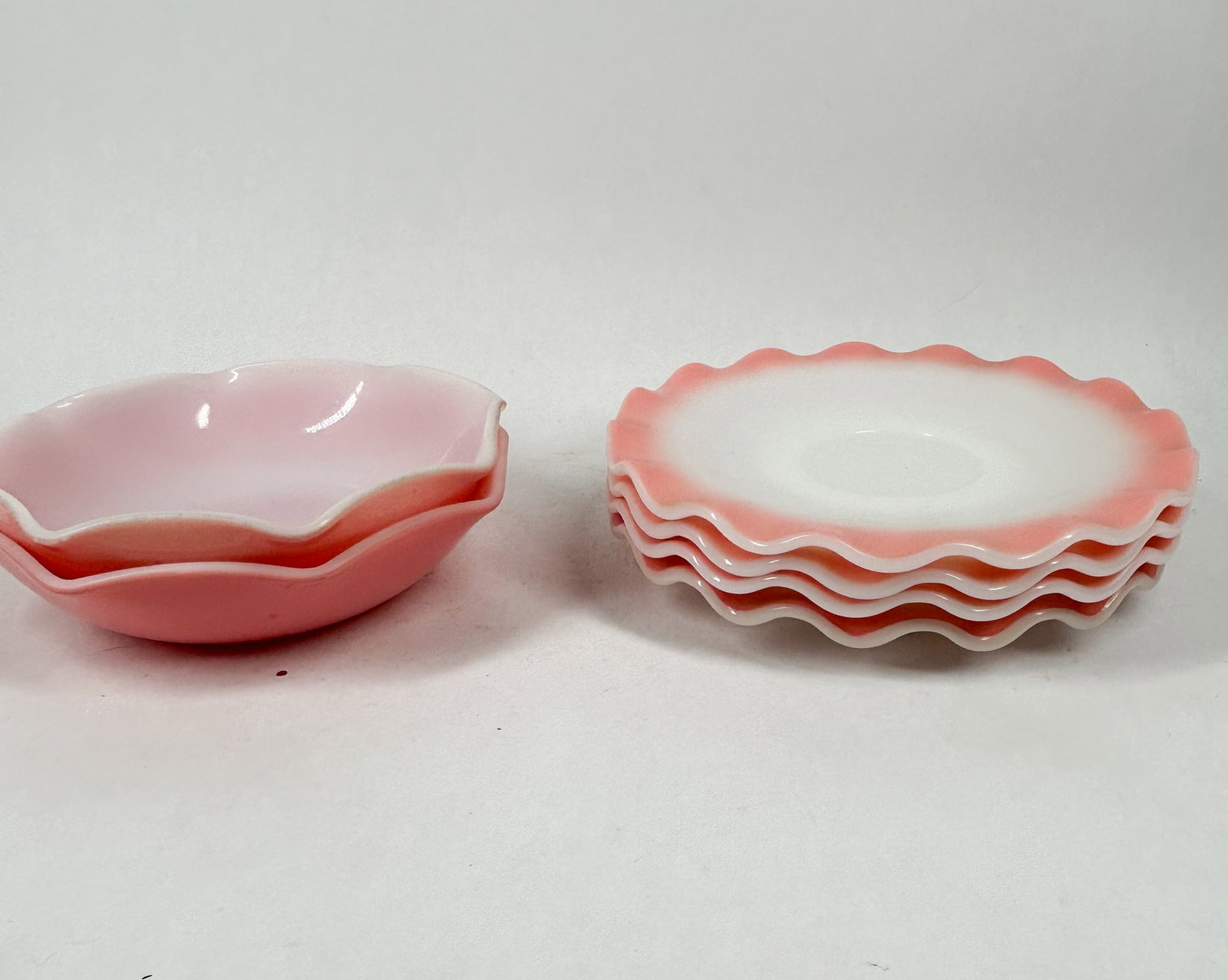 HAZEL ATLAS RUFFLED TABLEWARE: A collection of milk glass dessert bowls and saucers featuring scalloped rims and a fired-on pink gradient. An embossed monogram consisting of the letter H with a nested A is visible on the underside