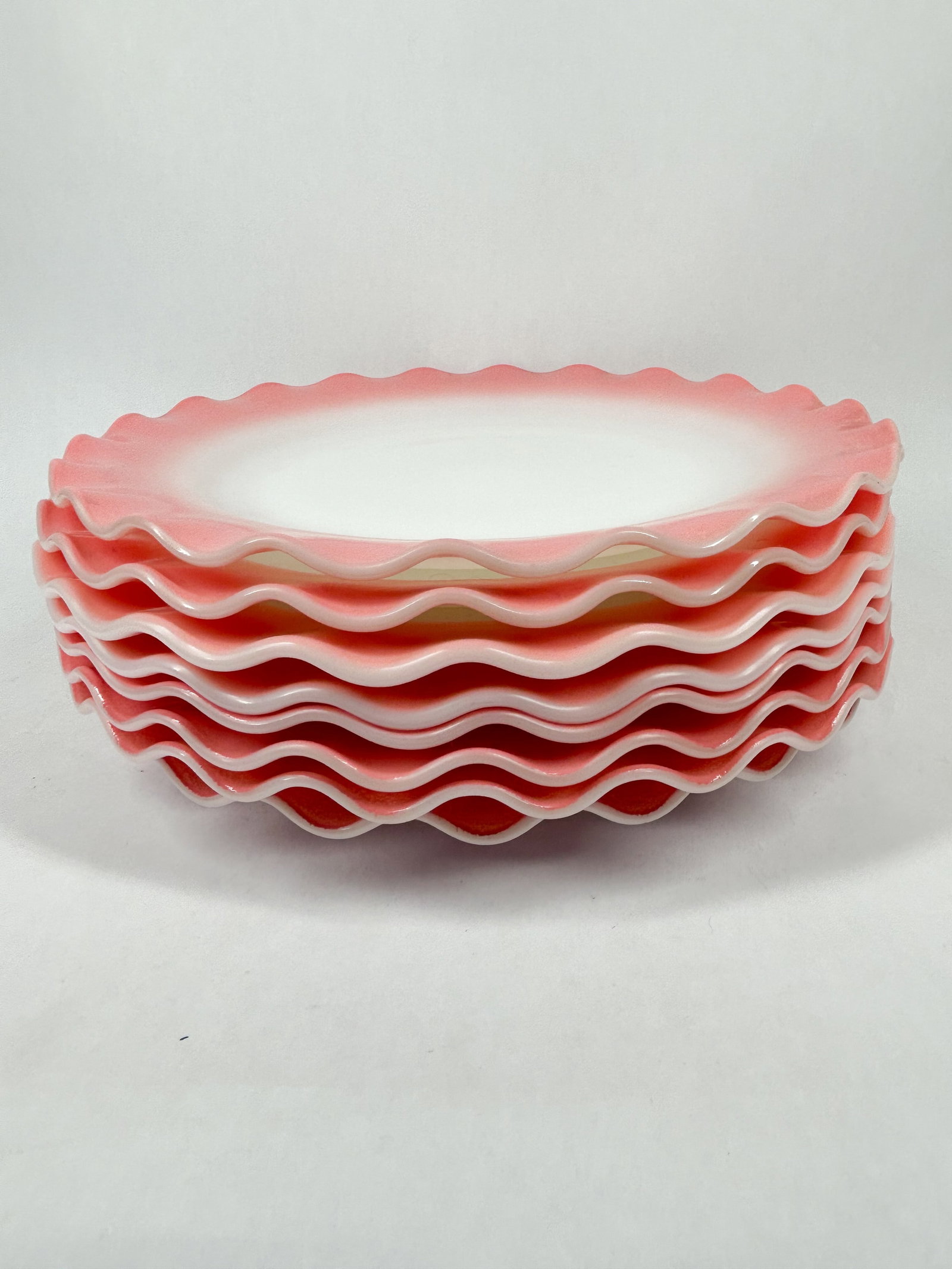 HAZEL ATLAS RUFFLED PLATES: A group of circular milk glass plates featuring fired-on pink gradient rims and ruffled edges. A small embossed character resembling a capital I with horizontal terminals is present in the center of