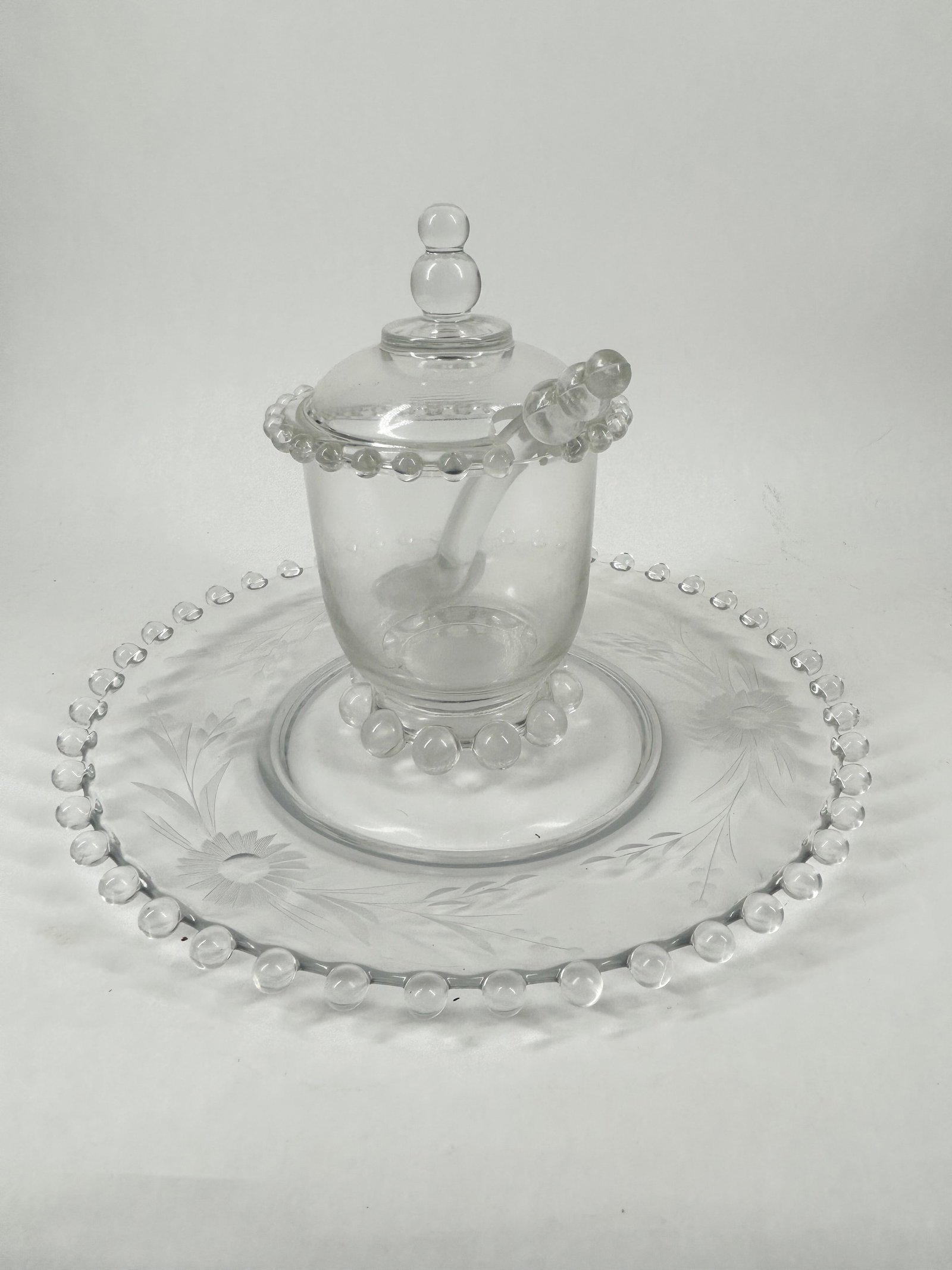 BEADED GLASS CONDIMENT SET, MID-20TH CENTURY: Three clear pressed glass items comprising a lidded jar, a spoon, and a tray. Each component is decorated with a repeating beaded border, and the tray features a delicate etched floral and foliate