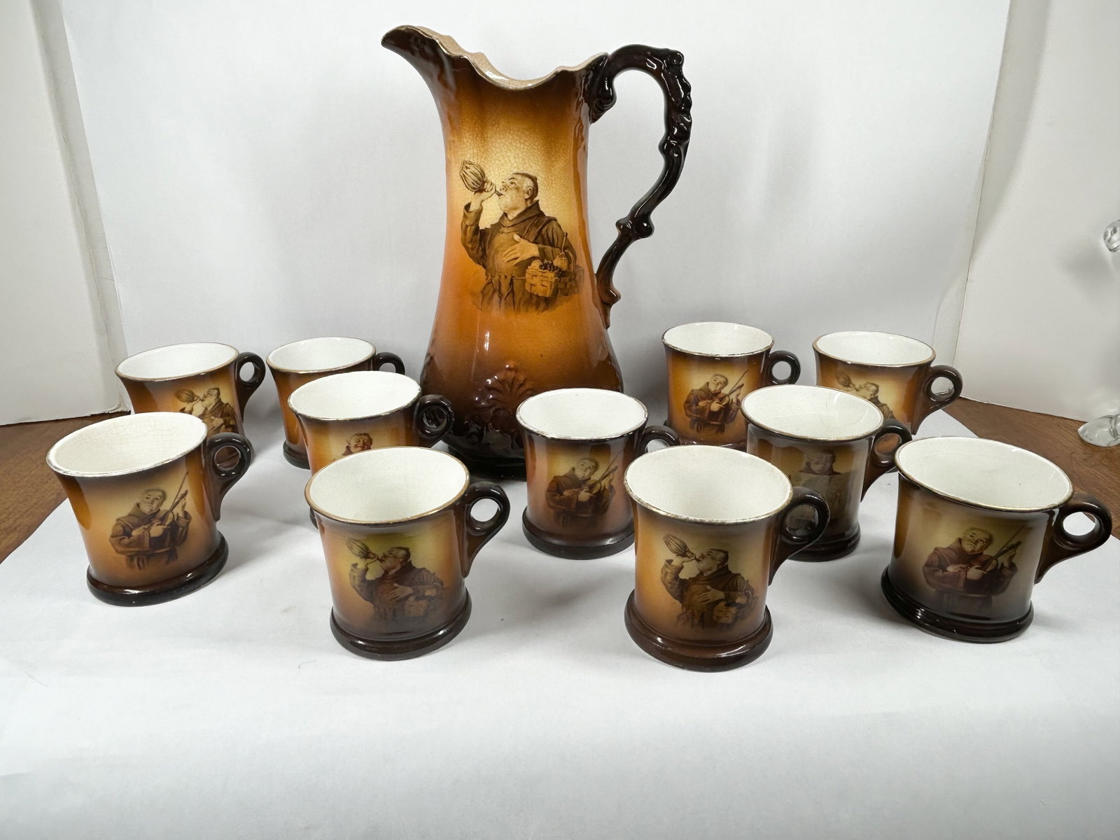 CERAMIC MONK PORTRAIT ALE SET: A group of ceramic ale vessels featuring transfer-printed monk vignettes and an amber-brown ombr? glazed finish. The set consists of a tall pitcher with a ruffled rim and scrolled handle along with
