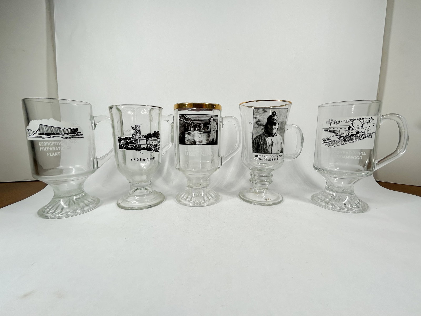 I.M.M.F. GLASS COMMEMORATIVE MINING MUGS, 1992-1997: A collection of I.M.M.F. clear glass footed commemorative mugs featuring graphics related to Ohio coal mining history, including the Georgetown Preparation Plant and Ida Mae Stull. The reverse of