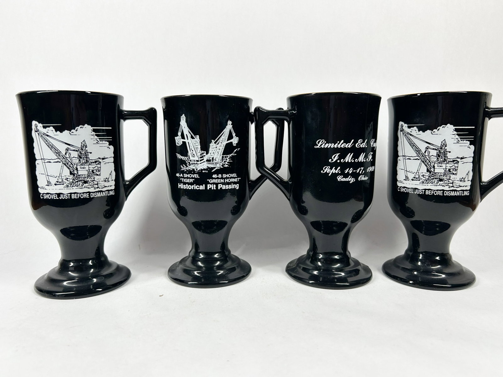 MINING MUGS AND CERAMIC TREE, 1989: A group of four black opaque glass footed mugs commemorating the 1989 Historical Pit Passing in Cadiz, Ohio, alongside a vintage green-glazed ceramic tabletop Christmas tree. The mugs feature white