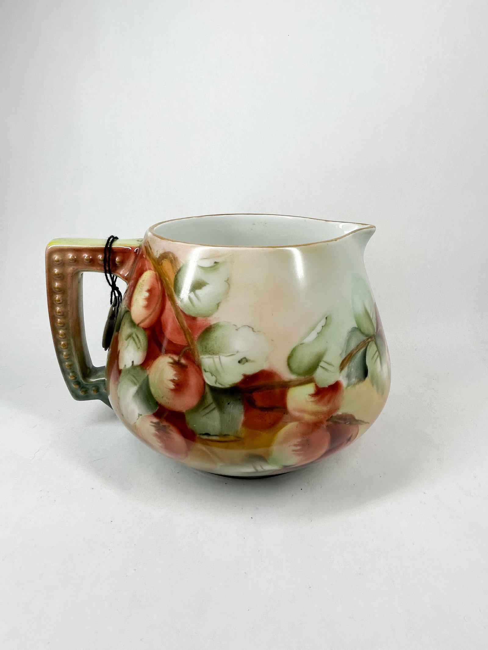 HAND PAINTED PORCELAIN CIDER PITCHER: Porcelain cider pitcher featuring a hand-painted motif of apples on leafy branches and a squared handle with gilded beading. A handwritten white string tag is attached to the handle reading JE-AT $68.