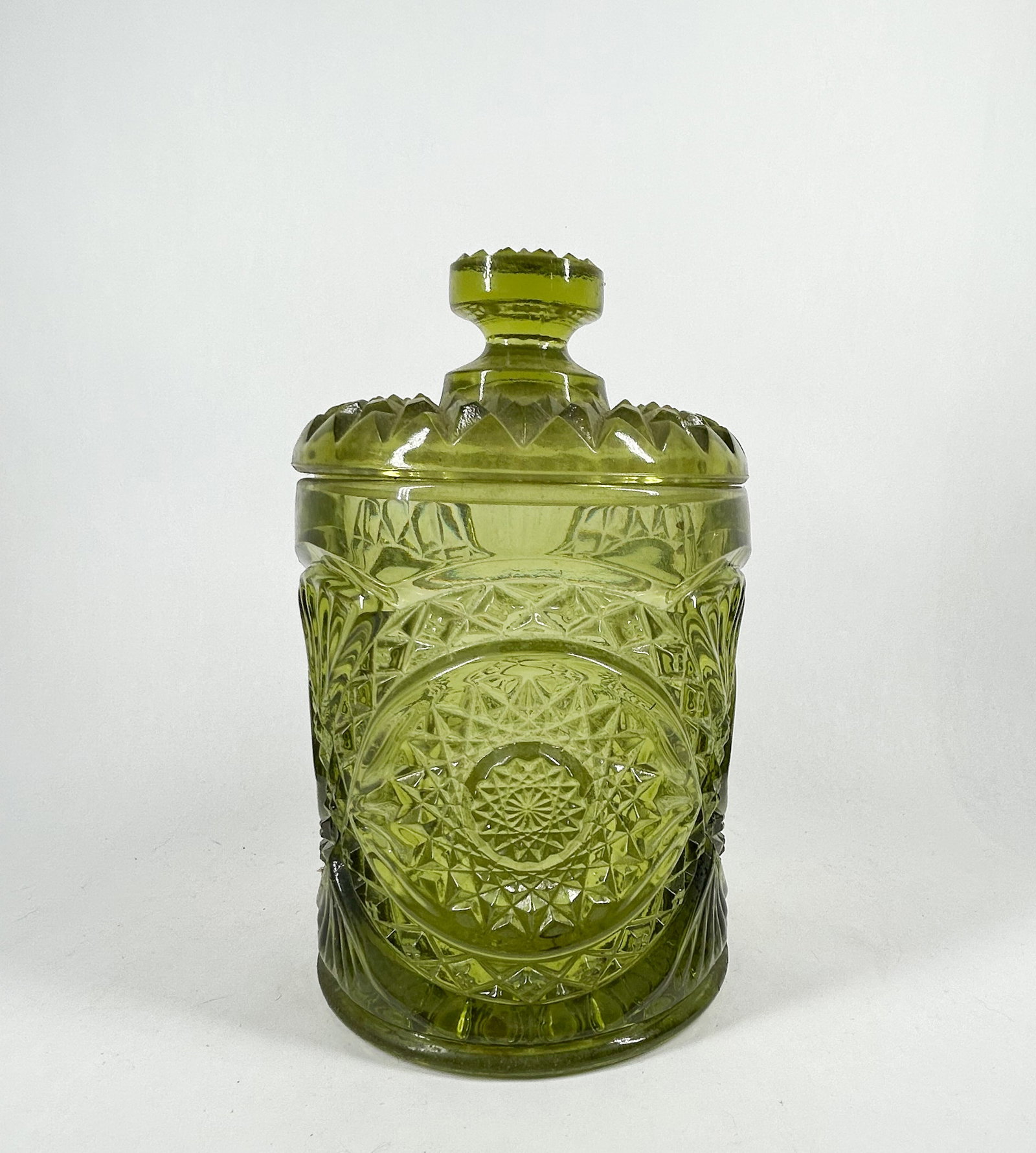 OLIVE GREEN PRESSED GLASS COVERED JAR, MID-20TH CENTURY: A lidded storage jar in translucent olive green pressed glass, featuring an all-over geometric pattern of hobstar medallions and fan-like fluting. A molded starburst is present on the center of the un
