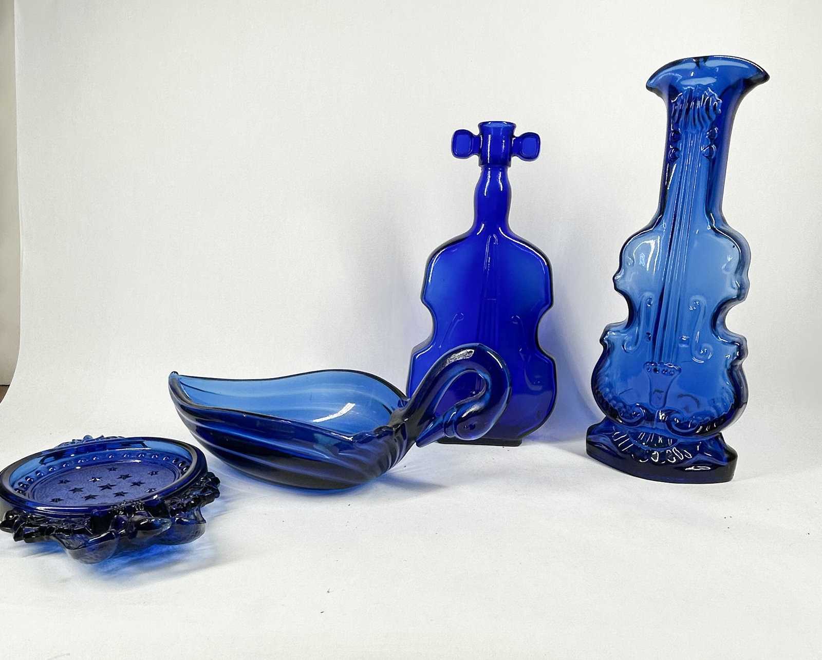 COBALT BLUE PRESSED GLASS COLLECTION, 20TH CENTURY: An assortment of cobalt blue pressed glass vessels featuring a patriotic eagle dish with stars, a ribbed swan-form bowl, a violin-shaped decanter with matching stopper, and a relief-molded violin vase