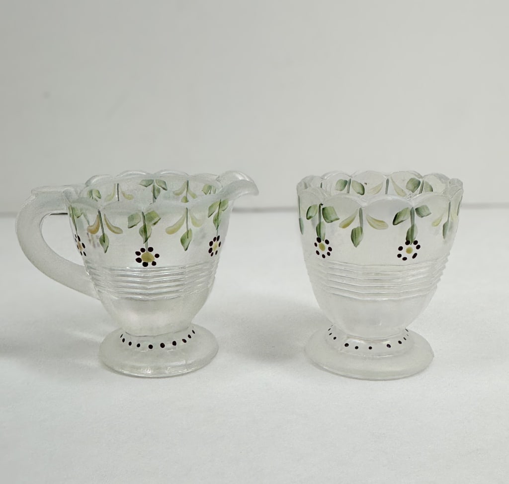 HAND-PAINTED FROSTED GLASS CREAMER AND SUGAR: A pair of frosted glass vessels comprising a creamer and an open sugar bowl, each featuring hand-painted floral motifs and molded horizontal ribbing. A molded sunburst pattern is visible on the unders