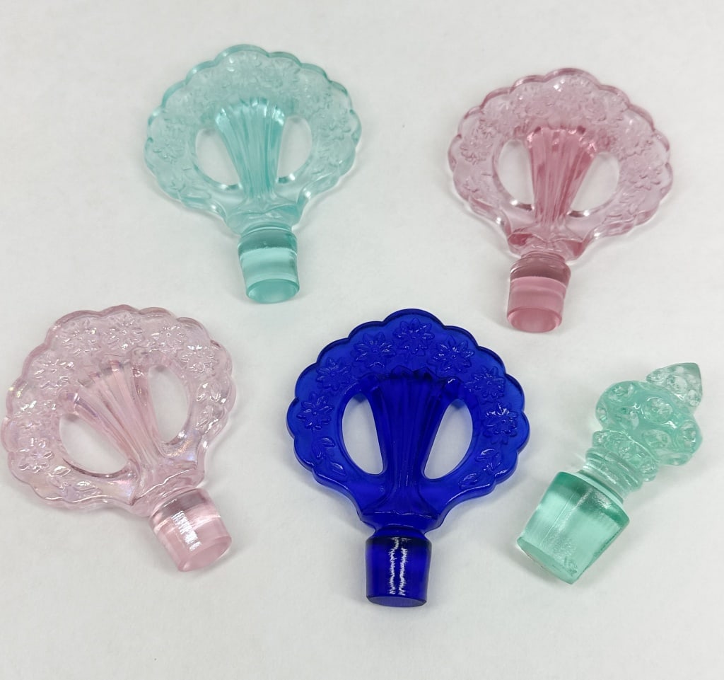 GLASS PERFUME STOPPERS, MID-20TH CENTURY: An assortment of pressed glass perfume bottle stoppers featuring floral openwork fan and finial designs. The collection includes translucent cobalt blue, pink, iridescent carnival glass, and aqua fini
