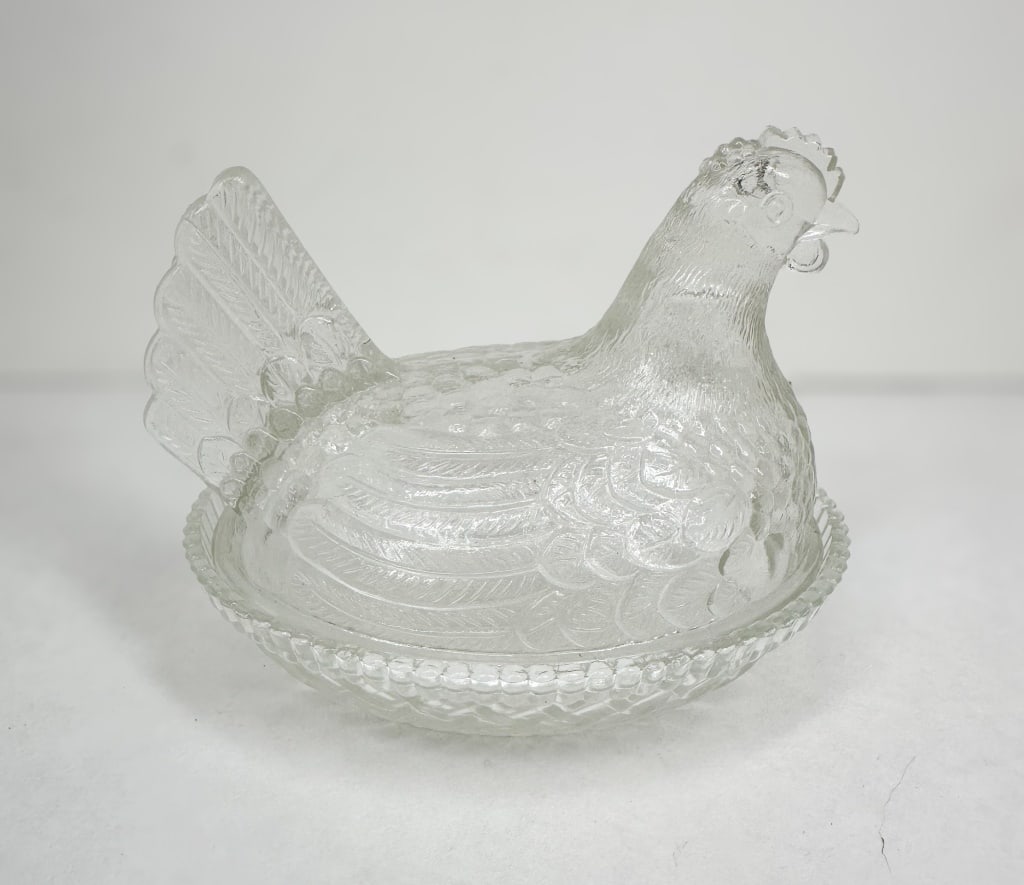 VINTAGE CLEAR GLASS HEN ON NEST: A two-piece vintage clear pressed glass covered dish in the form of a nesting hen. The object consists of an oval basket-weave base with a scalloped rim and a detailed lid featuring a flared tail and