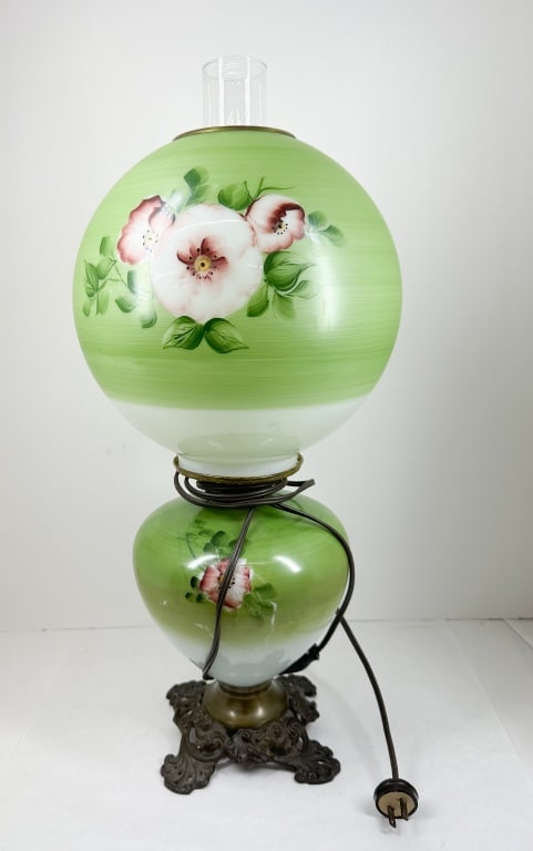 ELECTRIFIED GREEN FLORAL GLASS LAMP: Electrified parlor lamp with hand-painted floral sprays on a green and white gradient milk glass font and shade. The piece is mounted on an ornate four-footed cast metal base and features illegible