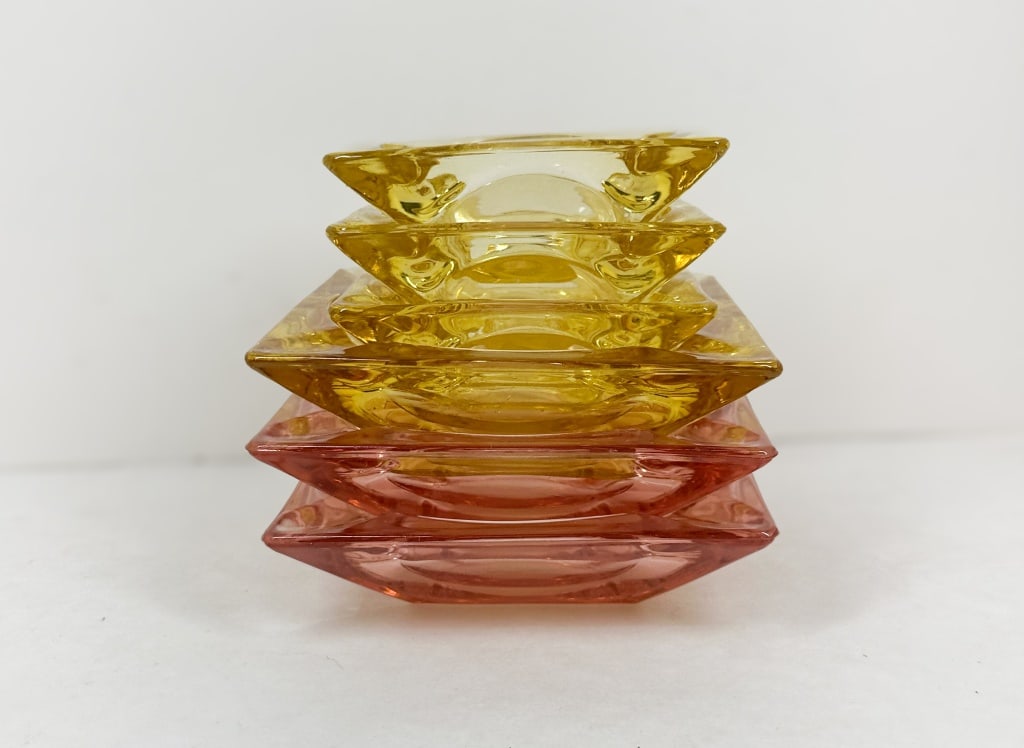 SIX MID-CENTURY PRESSED GLASS ASHTRAYS: A set of six vintage pressed glass ashtrays featuring square forms with circular wells and notched cigarette rests. The collection comprises amber and rose pink glass examples in two sizes.
