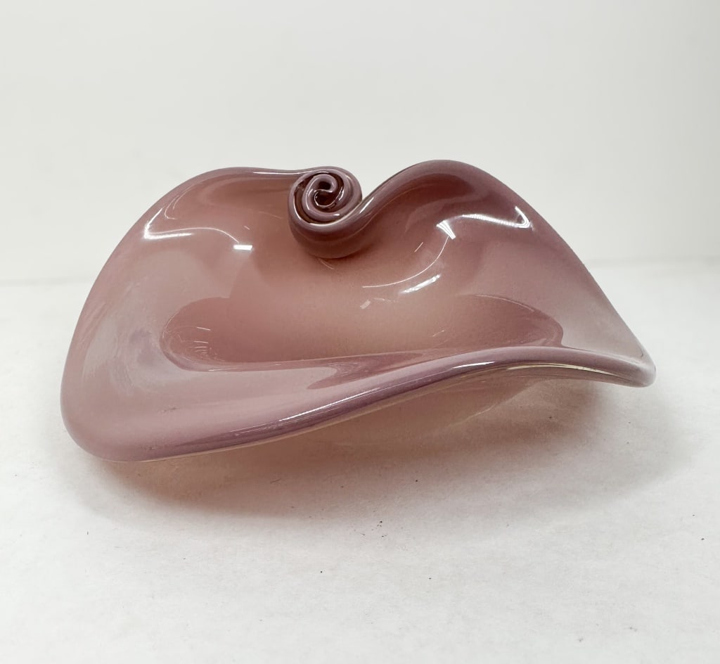 MID-CENTURY MAUVE ART GLASS BOWL: Hand-blown art glass bowl in a translucent mauve colorway featuring a freeform, shell-inspired design with an applied snail-coil spiral. A dark rectangular mark is visible on the ground and polished b