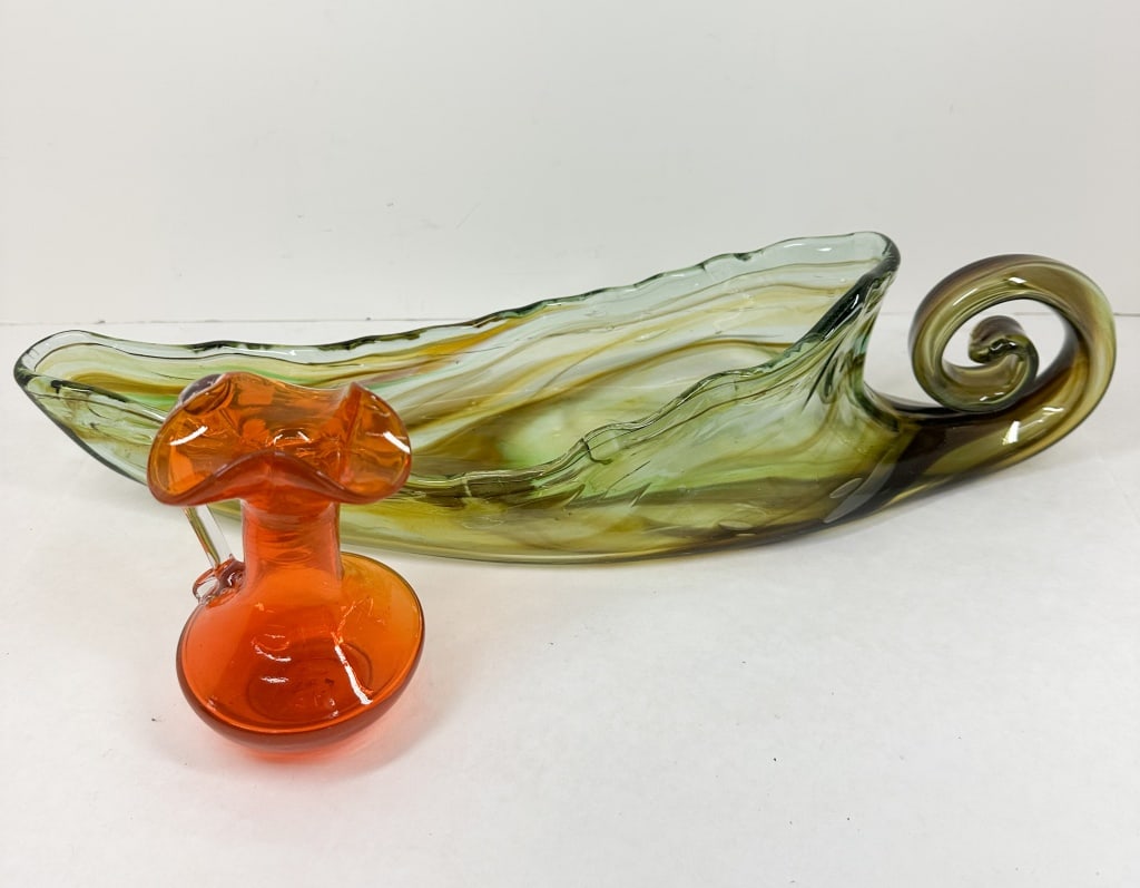 TWO PIECES HAND-BLOWN ART GLASS, MID-CENTURY: A collection of mid-century hand-blown art glass consisting of an elongated bowl with green and amber swirls and a scroll finial, and a translucent orange pitcher with a ruffled rim and clear applied 