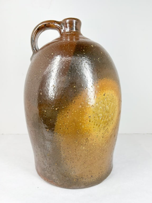 AMERICAN STONEWARE ALKALINE-GLAZED OVOID JUG: A 19th-century ovoid stoneware jug featuring a single pulled strap handle and a rich, mottled alkaline glaze. The surface displays characteristic iron spotting, pinhole pitting, and a prominent circul