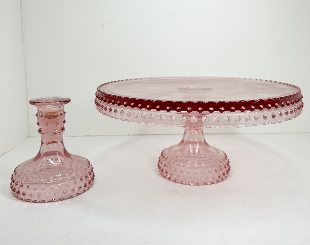 PINK HOBNAIL GLASS CAKE SET: A pink pressed glass cake stand and matching candlestick decorated in a hobnail pattern. The cake stand features a wide flat top with a ruby-stained beaded rim.