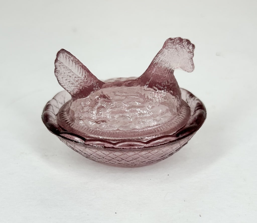 AMETHYST GLASS MINI HEN ON NEST: A two-piece covered pressed glass dish in the form of a nesting hen atop a basketweave base. The amethyst-colored glass features a textured finish on the hen's body and a scalloped rim on the base. A 