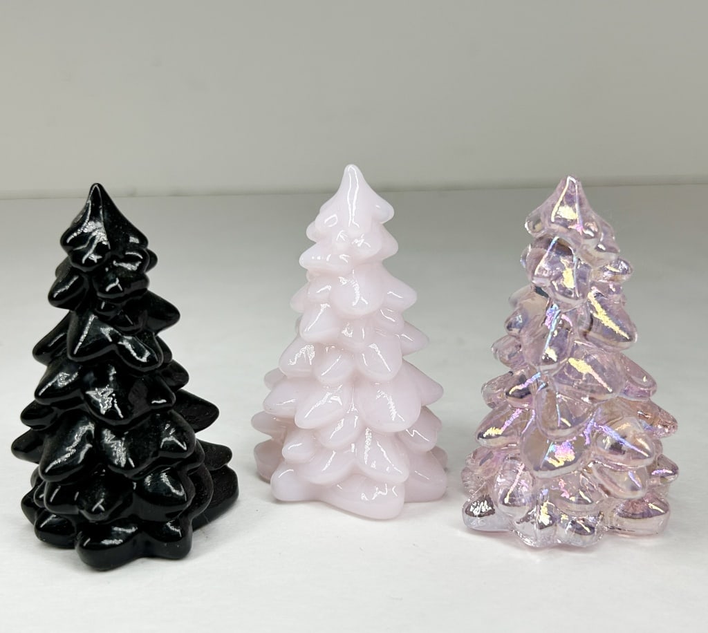 TRIO OF MINI MOSSER TREES: A set of three stylized evergreen figurines featuring tiered boughs and pointed tops. The collection includes one opaque black tree with a high-gloss finish, one opaque milk glass style pink tree, and