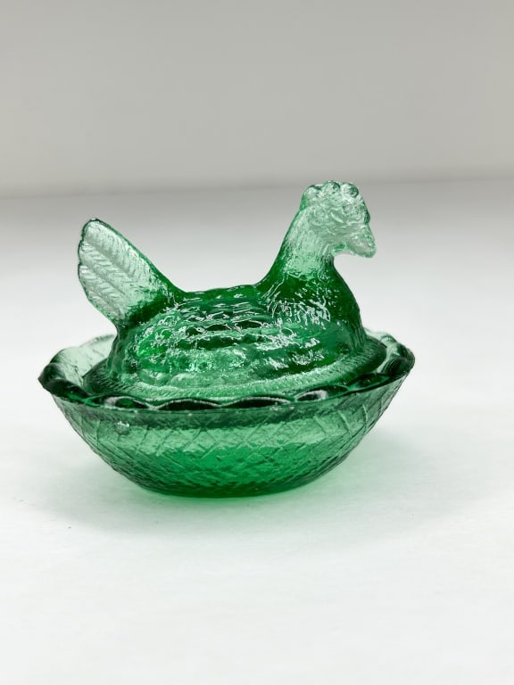 GREEN MINIATURE HEN ON NEST: A two-piece miniature lidded dish in transparent emerald green pressed glass. The lid is molded as a nesting hen with textured feathers and a split tail, resting upon a basketweave base with a scallop