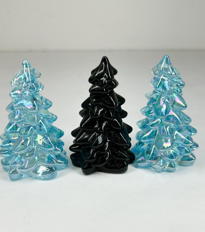 TRIO OF MOSSER GLASS CHRISTMAS TREES: A set of three contemporary stylized evergreen trees featuring tiered branch motifs. The group includes two trees crafted in iridescent blue carnival glass and one tree in opaque black polished glass.