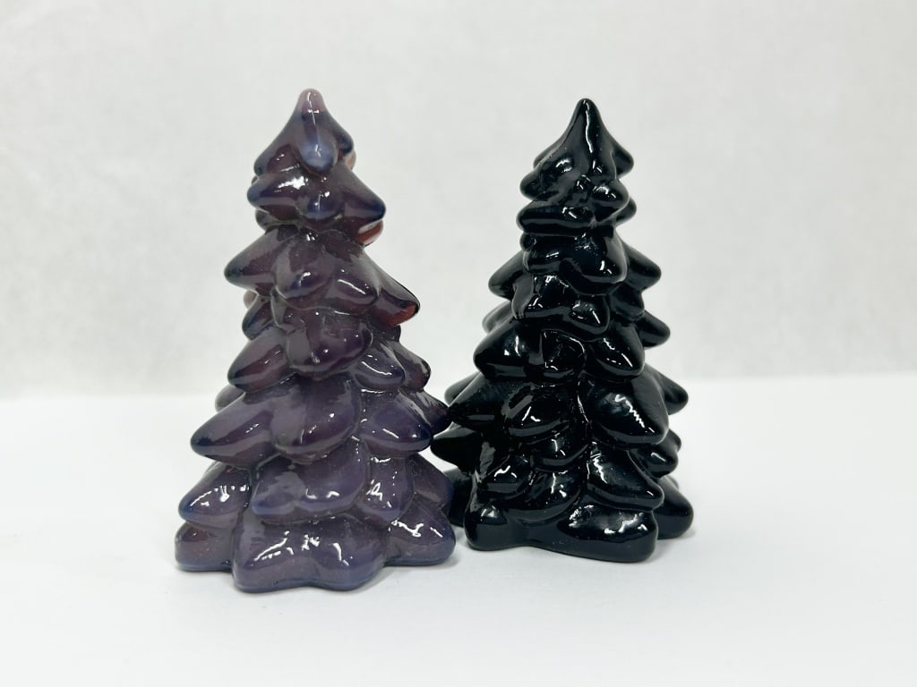 TWO MOSSER CHRISTMAS TREES: A pair of glass figurines in the form of stylized evergreen trees, featuring layered, rounded branches. One specimen is composed of semi-translucent purple slag glass with amethyst and lavender swirls