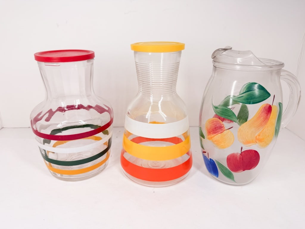 THREE GLASS JUICE CARAFES AND PITCHER: A trio of vintage glass vessels comprising two carafes and one pitcher. One carafe features a bulbous body with horizontal bands of maroon, green, white, and orange, fitted with a red plastic lid;