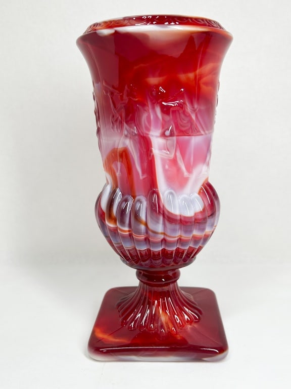 RED AND WHITE SLAG GLASS URN VASE: A vintage footed urn vase crafted in red and white marbled slag glass, featuring an embossed neoclassical frieze of draped female figures above a gadrooned bowl. The piece is supported by a ribbed ste